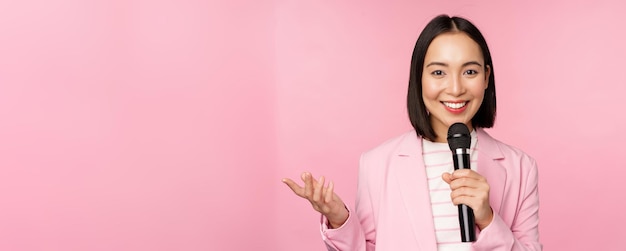 Mujer de negocios asiática dando un discurso sosteniendo el micrófono y sonriendo de pie en traje sobre fondo rosa Copiar espacio