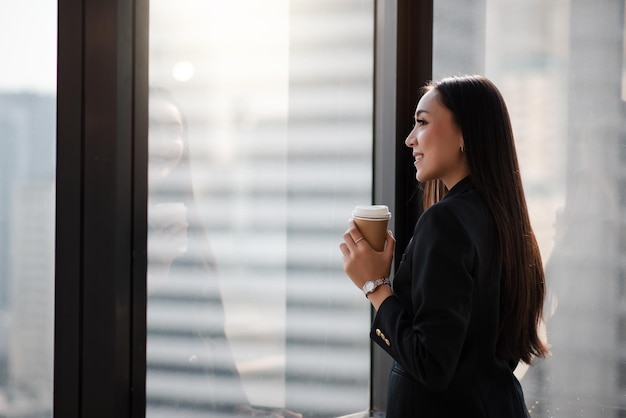 Mujer de negocios asiática inteligente adulta joven en traje casual negro sosteniendo una taza de café en el tiempo de descanso