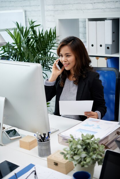 Mujer de negocios asiática sentada en la oficina, sosteniendo el documento y hablando por teléfono móvil