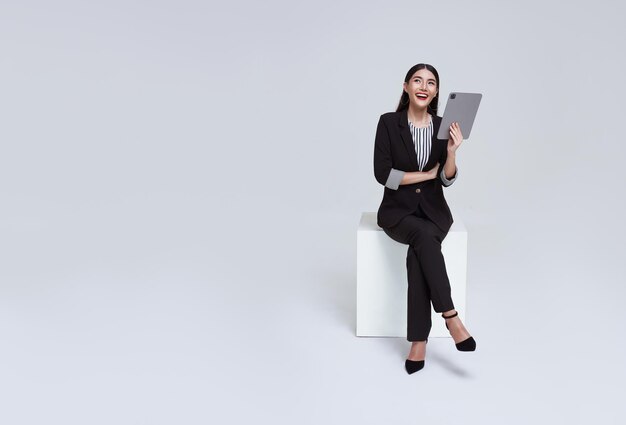 una mujer de negocios asiática sonríe con traje formal sentada en una silla usando una tableta y mirando al espacio de copias