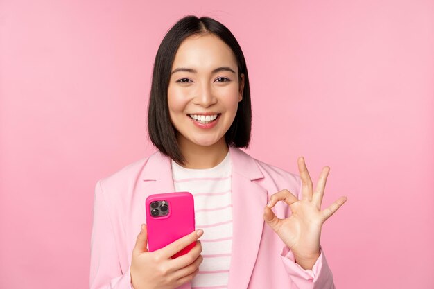 Mujer de negocios asiática sonriente que muestra un signo de aprobación mientras usa una aplicación de teléfono móvil recomendando una aplicación de teléfono inteligente de pie sobre un fondo rosa