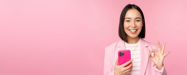 Mujer de negocios asiática sonriente que muestra un signo de aprobación mientras usa una aplicación de teléfono móvil recomendando una aplicación de teléfono inteligente de pie sobre un fondo rosa