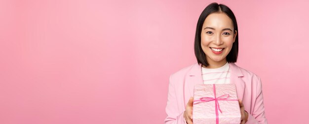 Mujer de negocios asiática sonriente en traje que le da un regalo en una caja envuelta de pie sobre fondo rosa