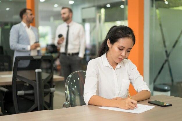 Mujer de negocios bastante asiática centrada escribiendo en el escritorio en la oficina