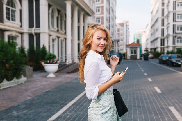 Mujer de negocios bastante rubia con el pelo largo en camisa blanca caminando por la calle alrededor del barrio británico. Ella sostiene el teléfono