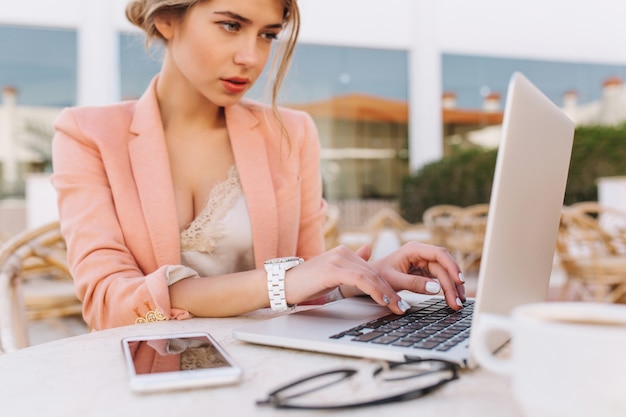 Mujer de negocios bonita joven que trabaja en la computadora portátil en el café de la calle, escribiendo en el teclado, señora inteligente que mira seriamente en la pantalla. smartphone y gafas en la mesa. vistiendo elegante chaqueta rosa, relojes blancos.