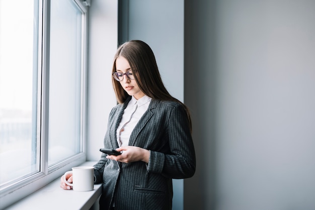 Mujer de negocios con café usando teléfono inteligente en la ventana