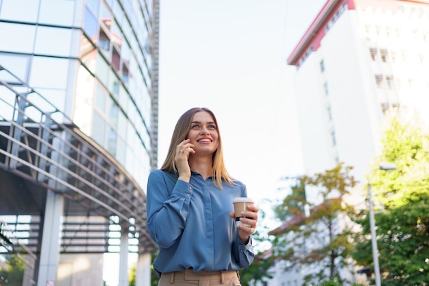 Mujer de negocios caucásica hablando por teléfono con café para llevar. Una mujer europea exitosa, hablando por teléfono, de pie en el edificio de oficinas moderno