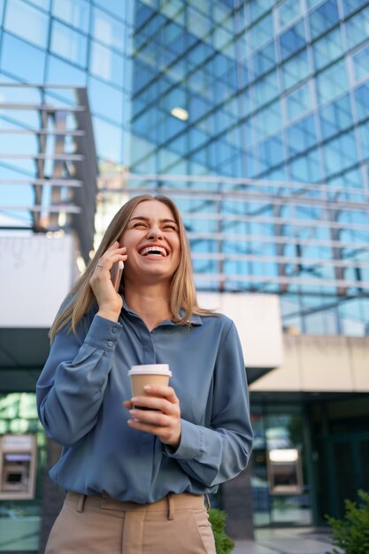 Mujer de negocios caucásica hablando por teléfono con café para llevar. Una mujer europea exitosa, hablando por teléfono, de pie en el edificio de oficinas moderno