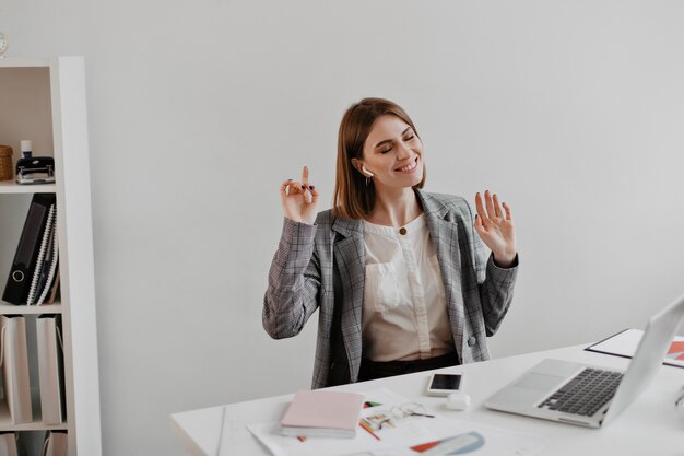 Mujer de negocios en chaqueta gris disfrutando de la música mientras está sentado en el lugar de trabajo en la oficina blanca.