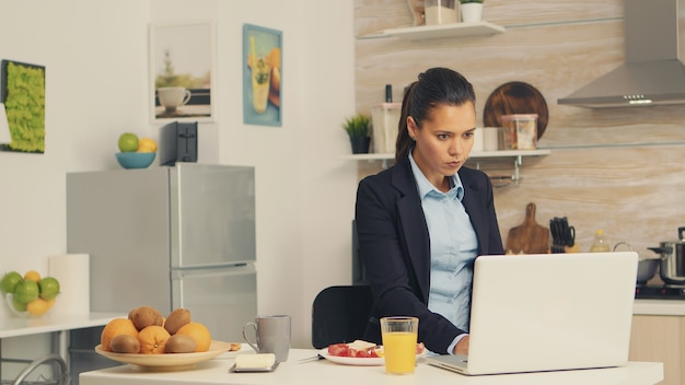 Mujer de negocios comiendo pan tostado con mantequilla mientras trabaja en la computadora portátil durante el desayuno. Mujer de negocios concentrada en la mañana multitarea en la cocina antes de ir a la oficina, estresante w