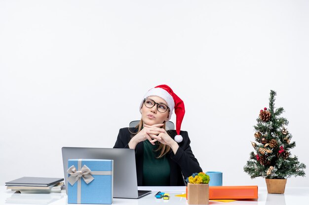 Mujer de negocios concentrada positiva con su sombrero de santa claus sentado en una mesa con un árbol de Navidad y un regalo y pensando en algo sobre fondo blanco