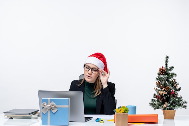 Foto gratuita mujer de negocios concentrada con un sombrero de santa claus sentado en una mesa con un árbol de navidad y un regalo y revisando sus correos sobre fondo blanco.