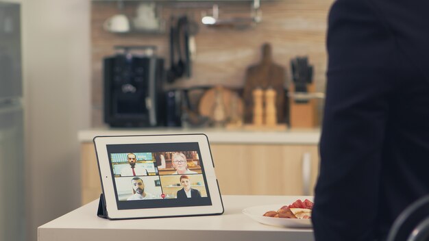 Mujer de negocios con dispositivo inteligente mientras desayuna en la cocina. Joven autónomo en casa hablando en una videollamada con sus colegas de la oficina, utilizando tecnología moderna de Internet