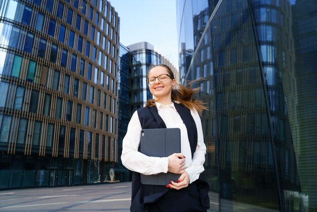 Una mujer de negocios se encuentra con una computadora portátil en un traje y gafas fuera de un edificio de oficinas durante el día.
