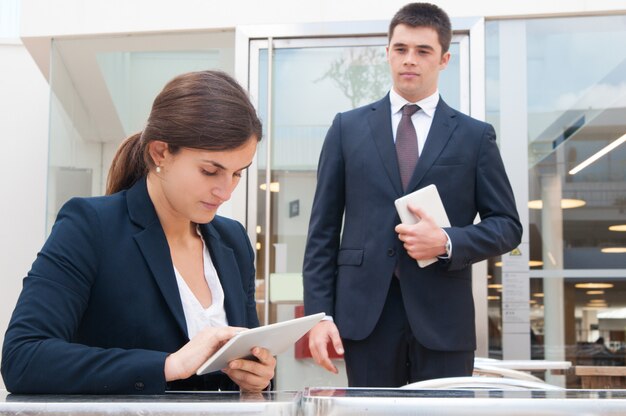 Mujer de negocios enfocada que usa la tableta y el compañero de trabajo que se colocan cerca