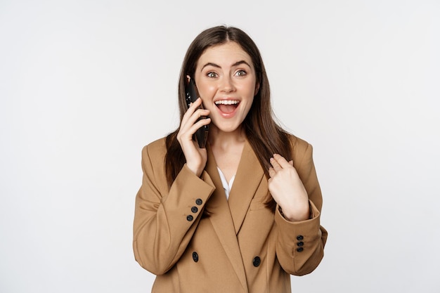 Mujer de negocios entusiasta hablando por teléfono móvil, reaccionando sorprendida y feliz de llamar, recibir buenas noticias, de pie sobre fondo blanco