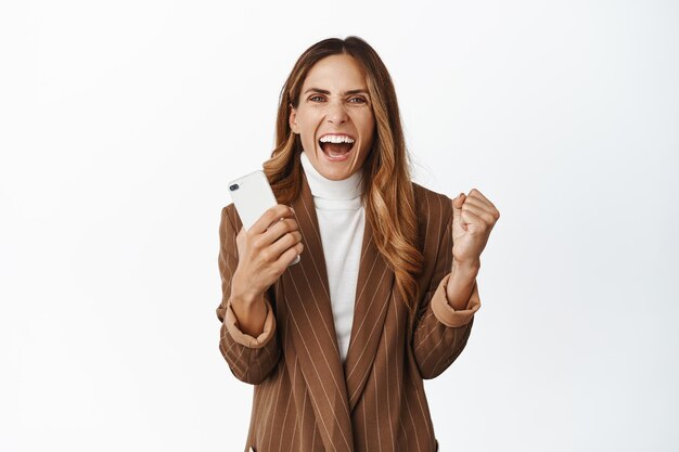 Una mujer de negocios entusiasta que gana en un teléfono inteligente logra el objetivo en una aplicación de teléfono móvil y grita emocionada celebrando de pie sobre un fondo blanco