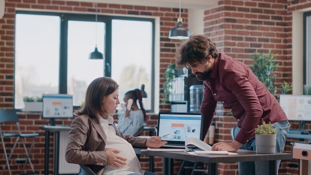 Mujer de negocios esperando un hijo y teniendo una discusión con el hombre mientras trabaja en la planificación del proyecto. Emprendedora embarazada hablando con un colega sobre análisis en archivos y computadora.