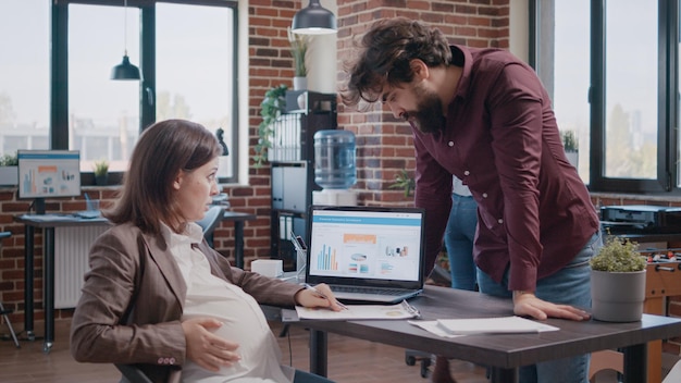 Mujer de negocios esperando un hijo, trabajando en análisis con un colega para la planificación del proyecto. Hombre ayudando a la empleada embarazada con la estrategia de crecimiento, desarrollo y marketing en la computadora portátil.