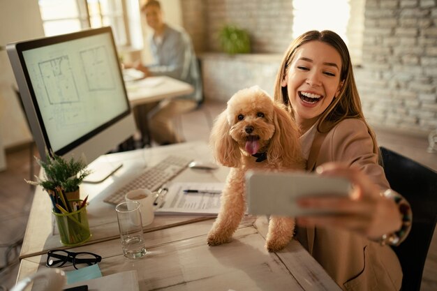 Mujer de negocios feliz divirtiéndose mientras toma selfie con su perro en la oficina