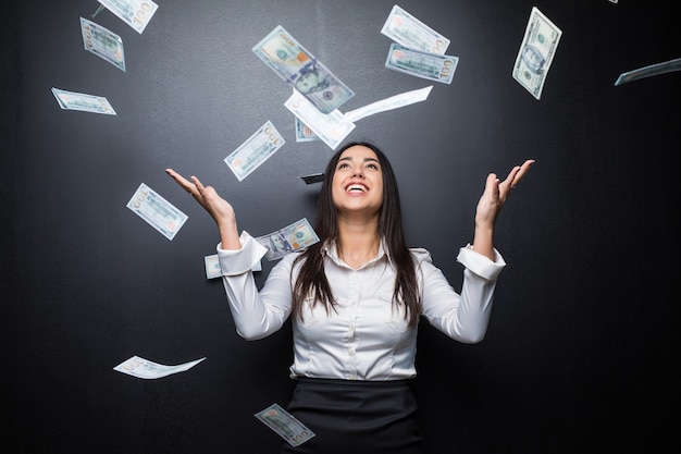Mujer de negocios feliz bajo una lluvia de dinero hecha de dólares aislado en la pared negra