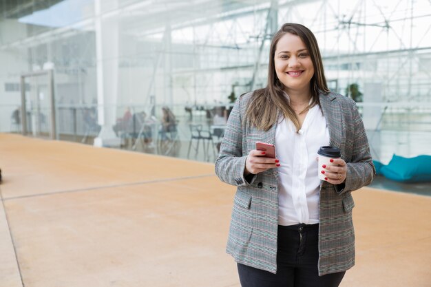 Mujer de negocios feliz que sostiene smartphone y la bebida al aire libre