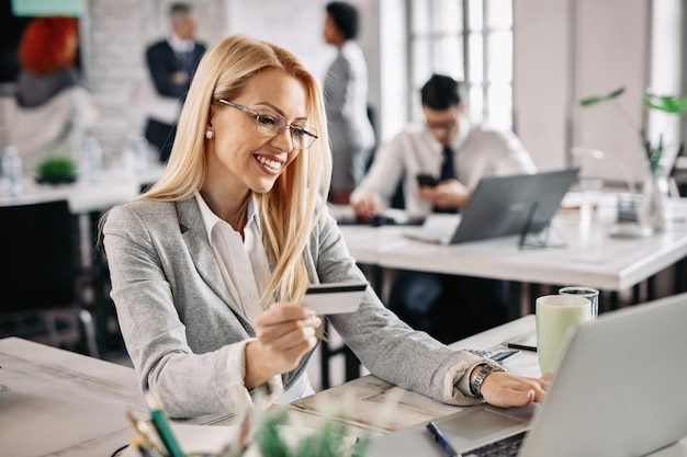 Mujer de negocios feliz que usa una computadora portátil y una tarjeta de crédito para comprar en línea mientras está sentada en su escritorio en la oficina