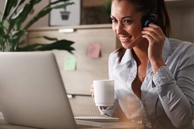 Mujer de negocios feliz tomando café mientras trabaja en el centro de atención al cliente y habla con un cliente por teléfono