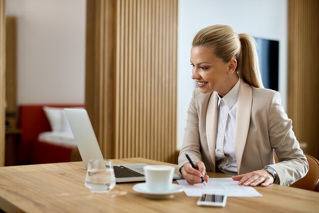 Mujer de negocios feliz tomando notas mientras trabaja en una computadora en la habitación del hotel