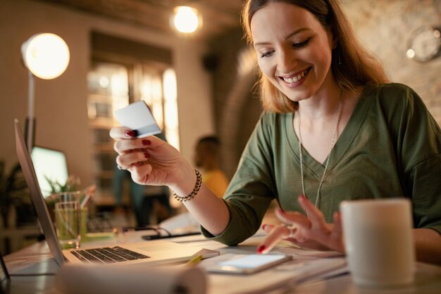 Mujer de negocios feliz usando teléfono inteligente y tarjeta de crédito mientras compra en Internet por la noche en la oficina