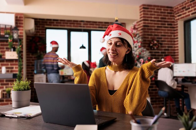 Mujer de negocios hablando en videoconferencia en la oficina con árbol de navidad y decoraciones festivas. Asistir a llamadas de teleconferencia remotas y reuniones en línea durante la temporada de vacaciones en el trabajo.