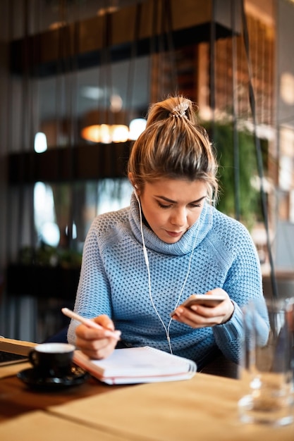 Mujer de negocios informal que trabaja en un café y toma notas mientras lee algo en el teléfono móvil