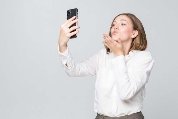 Mujer de negocios joven beso de golpe mientras toma selfie en el teléfono aislado en la pared blanca