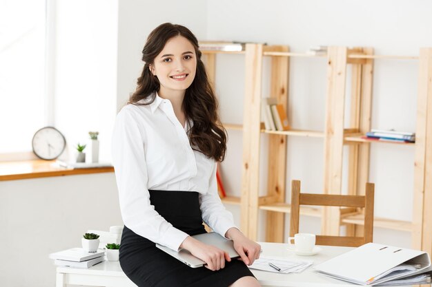 Mujer de negocios joven feliz o secretaria que sostiene la computadora portátil en la oficina moderna