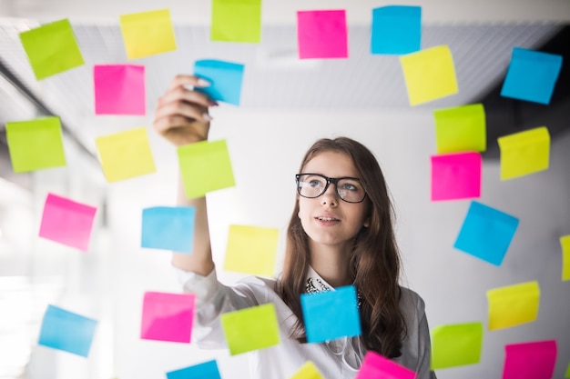 Mujer de negocios joven en gafas ver en pared transparente con un montón de pegatinas de papel en él