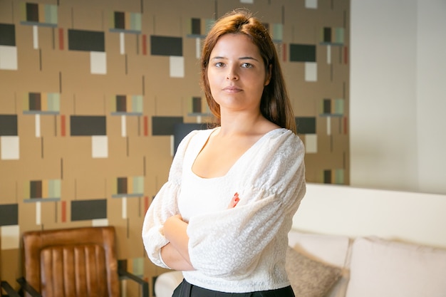 Mujer de negocios joven hermosa confiada que se coloca con los brazos cruzados y que presenta en el trabajo compartido o el interior de la cafetería, mirando a la cámara