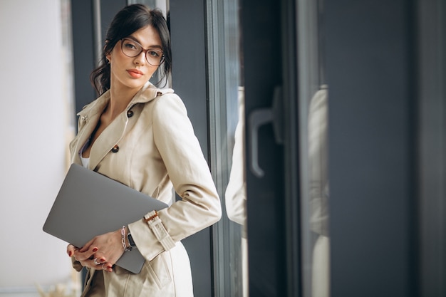 Mujer de negocios joven con laptop de pie junto a la ventana en la oficina