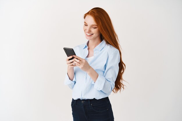 Mujer de negocios joven leyendo un mensaje en el teléfono inteligente y sonriendo, redes sociales mientras está de pie sobre una pared blanca