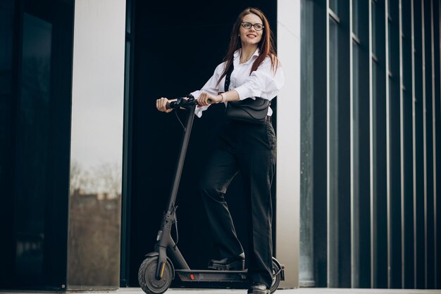 Mujer de negocios joven montando scooter eléctrico por centro de oficina