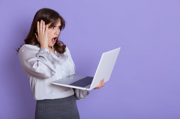 Mujer de negocios joven nerviosa atractiva con la computadora portátil en las manos, mirando la pantalla del dispositivo con expresión de asombro, tocando su cabeza con la mano, mantiene la boca abierta, se encuentra contra la pared lila.