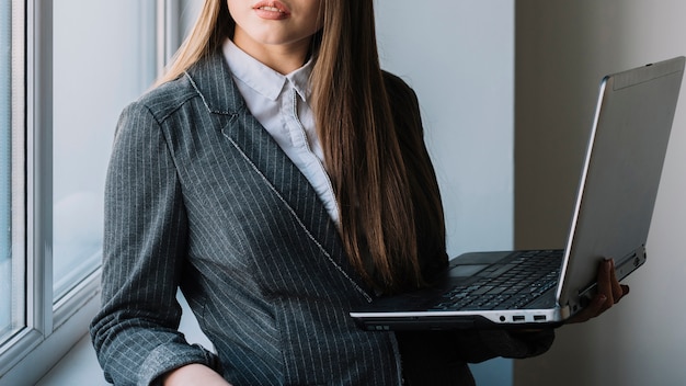 Mujer de negocios joven que se coloca en la ventana con la computadora portátil