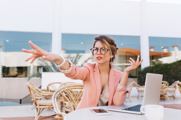 Mujer de negocios joven sentada en un café al aire libre con un portátil en la mesa, señora seria señalando con la dirección de la mano, vio algo en un afait. Vistiendo elegante chaqueta rosa, gafas, relojes blancos.