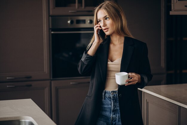 Mujer de negocios joven tomando café y hablando por teléfono en la cocina