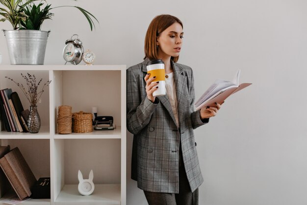 Mujer de negocios joven con un vaso de café en sus manos, fascinada por la lectura, se encuentra apoyada en el estante con accesorios de trabajo.