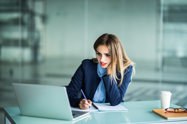 Mujer de negocios joven en vasos sentado en la mesa de madera con computadora portátil, planta, taza de café desechable y escribiendo en el papel, sosteniendo una pluma y un teléfono inteligente