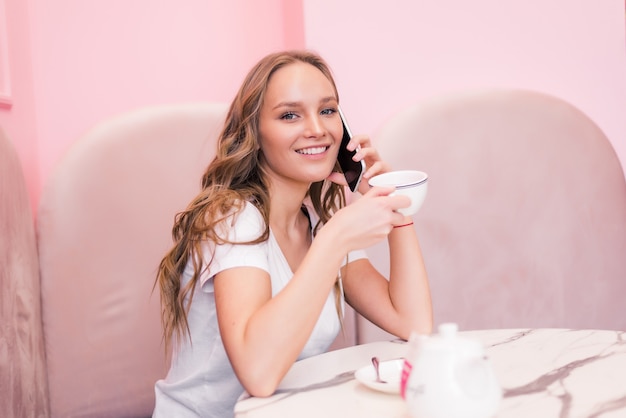Mujer de negocios joven en vestido gris sentado a la mesa en el café, hablando una vez por teléfono mientras toma notas en el cuaderno
