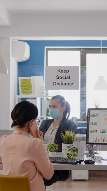 Mujer de negocios con mascarilla médica hablando por teléfono inteligente con socios mientras sus compañeros de trabajo trabajan ...