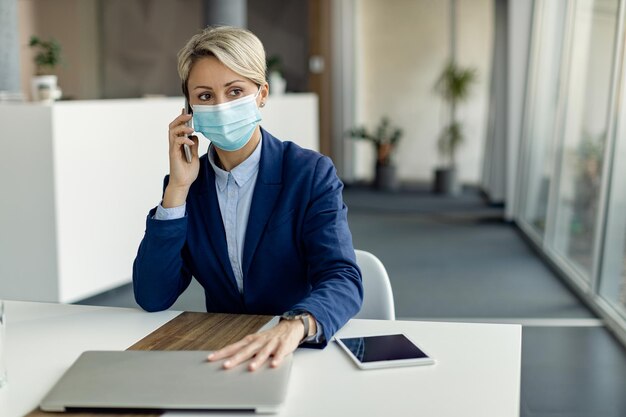 Mujer de negocios con mascarilla protectora hablando por teléfono celular mientras trabaja en la oficina