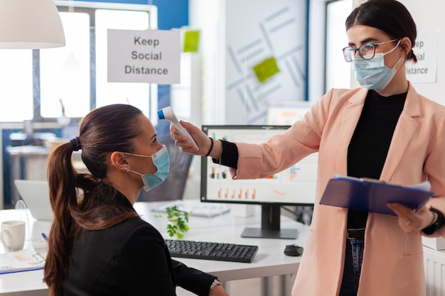 Mujer de negocios con mascarilla que toma la temperatura corporal de un colega en la oficina de la empresa usando un termómetro digital con infrarrojos, durante la pandemia global con coronavirus, manteniendo el distanciamiento social.